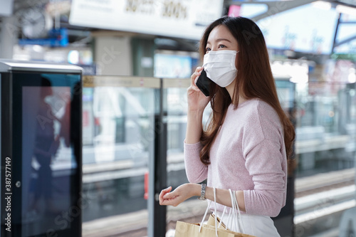 Asian business woman in casual dress code wearing face mask talking on mobile phone. She is waiting for the train to go to work on the platform station. New normal lifestyle in city concept.