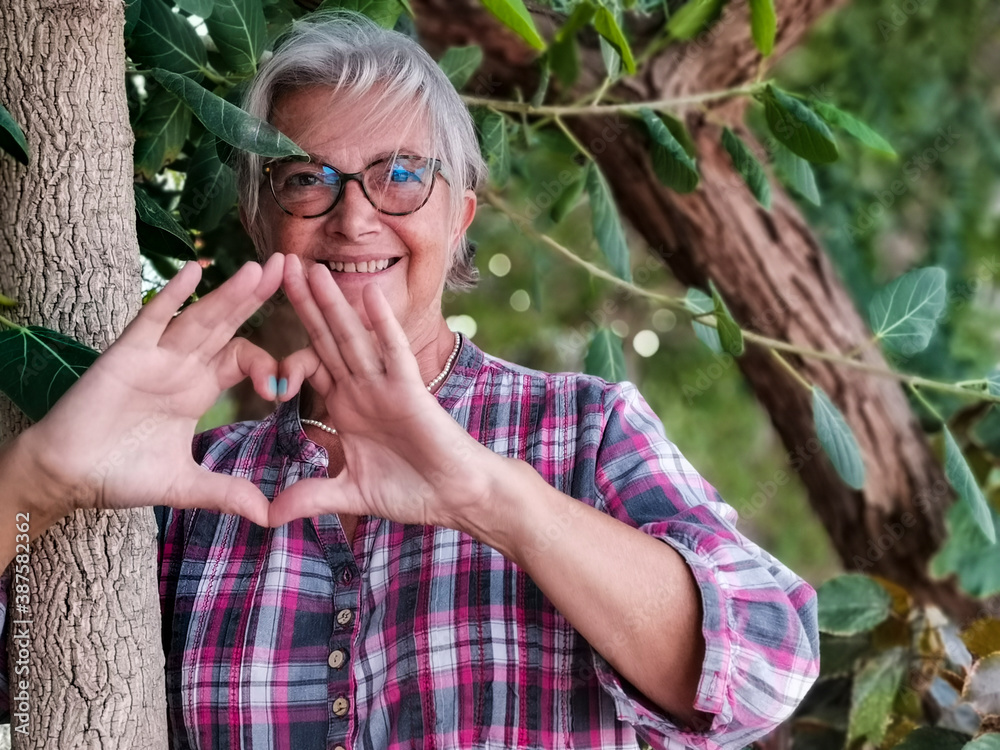 A senior smiling white haired woman enjoying nature amidst trees hugging a tree trunk making a heart shape with her hands. Active retired people who send love messages for nature A senior smiling white haired woman enjoying nature amidst trees hugging a tree trunk making a heart shape with her hands. Active retired people who send love messages for nature