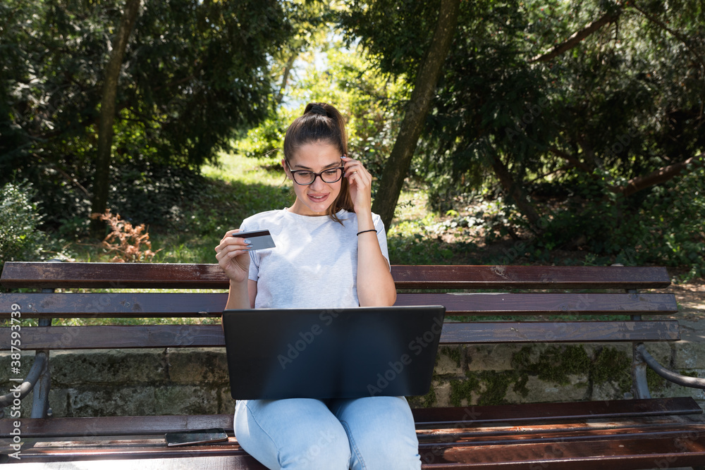 Obraz premium Happy young woman or girl sitting in the park with laptop and bank credit card and smiling after successful online shopping 