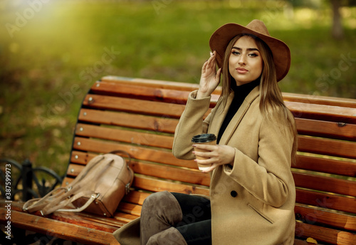 Young attractive woman in autumn clothes sits on bench and drinks coffee in city park. Woman dressed in stylish coat and felt hat. Autumn time
