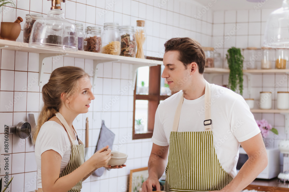 Fototapeta premium Happy & smiling attractive young cute caucasian couple in love enjoying cooking healthy salad in kitchen at home together. Happiness family of beautiful romantic married lover or marriage relationship