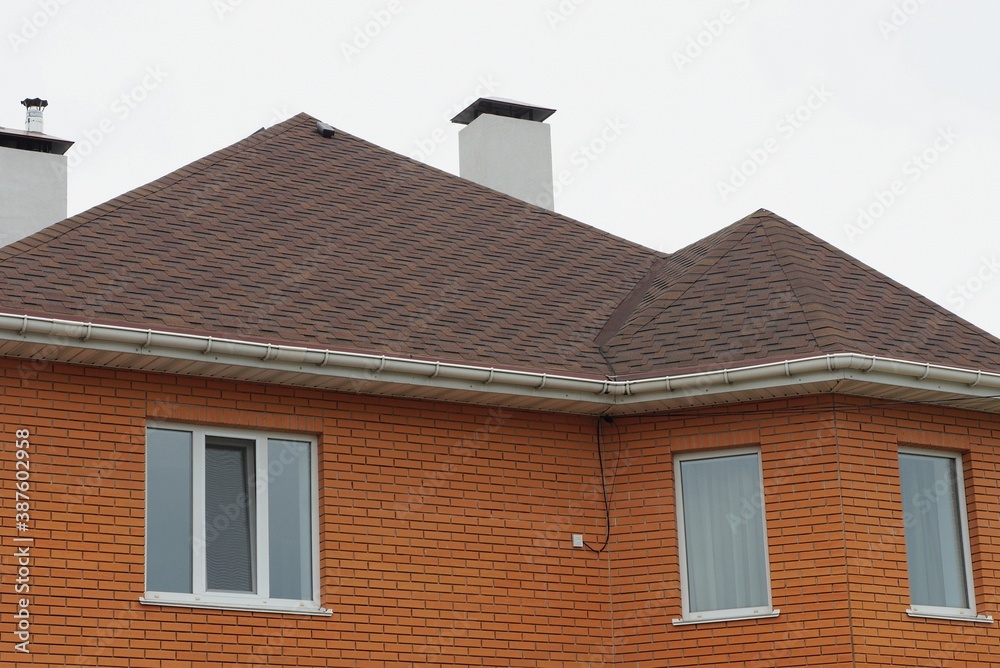 brick house with white windows and brown tiled roof with chimneys against gray sky brick house with white windows and brown tiled roof with chimneys against gray sky