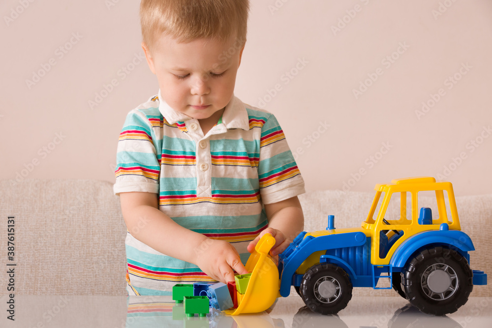 A blond boy in striped football plays at home in the children's room with colorful cubes and a large bright tractor on the table. A blond boy in striped football plays at home in the children's room with colorful cubes and a large bright tractor on the table.