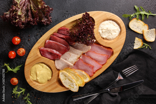 Snacks and appetizers. Meat cuts on a wooden plate with horseradish and croutons on a black table. Fresh lettuce, arugula and tomatoes. Restaurant menu. Background image, copy space. Flatlay, top view