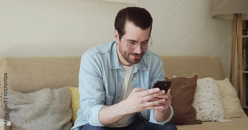 Relaxed young man in glasses sitting on comfortable couch, spending free weekend leisure time online, using smartphone applications, dating or communicating distantly with friends in social network. Relaxed young man in glasses sitting on comfortable couch, spending free weekend leisure time online, using smartphone applications, dating or communicating distantly with friends in social network.