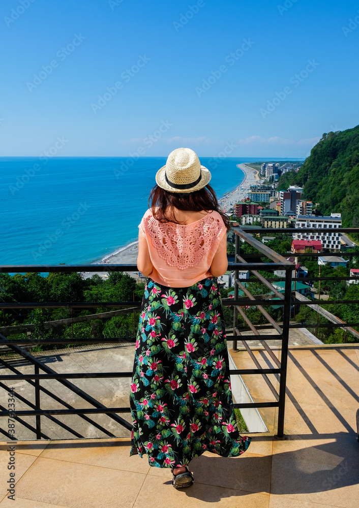 Girl portrait in straw hat, coral blouses and tropical print skirt. Looking on sea and panorama city. Back view. Young woman stands on balcony. Warm summer day and vacation time. Kvariati town