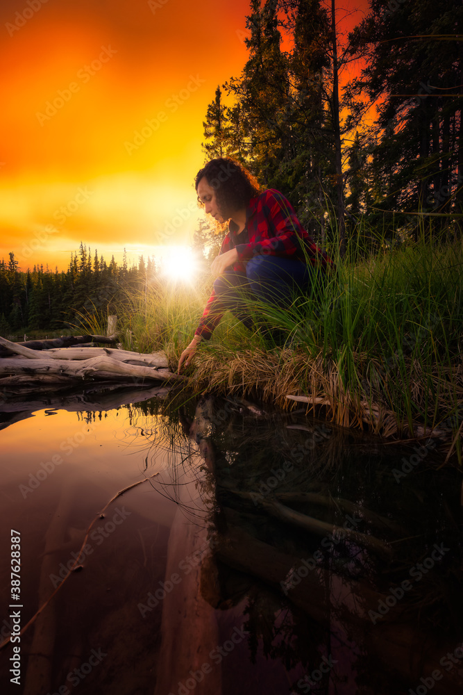 Girl Crouched by a Scenic Lake Reflecting on Life in Canadian Nature. Dramatic Colorful Sunset Artistic Render. Taken near Whitehorse, Yukon, Canada.