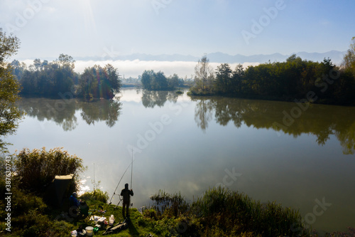 Fisherman fishing on the edge of the wild lake in beautiful morning light. Fishing and hunting concept. Catching fish on the lake among the green trees of the forest. Water reflection in soft light. 