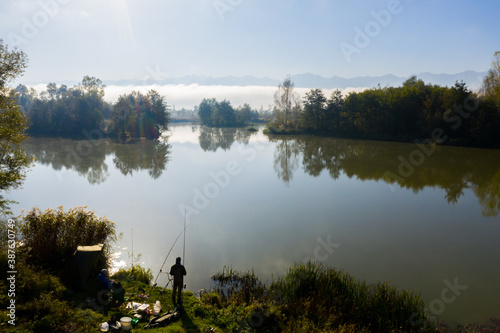 Fisherman fishing on the edge of the wild lake in beautiful morning light. Fishing and hunting concept. Catching fish on the lake among the green trees of the forest. Water reflection in soft light. 