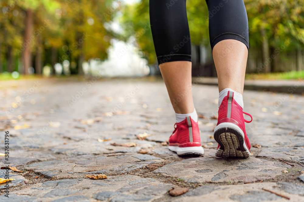 Young fitness a woman in sports red sneakers and black leggings walks in the park along a stone road in cloudy autumn weather. Sport, healthy and wellness concepts