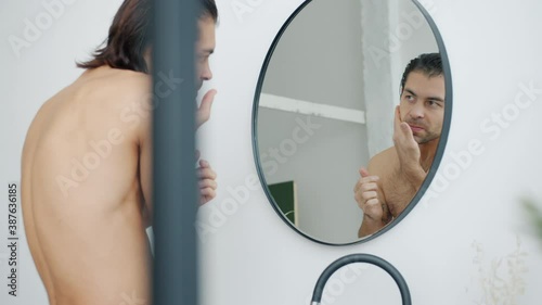 Good-looking young man is applying aftershave cream on face looking at mirror in bathroom caring for skin health. People and beauty concept.