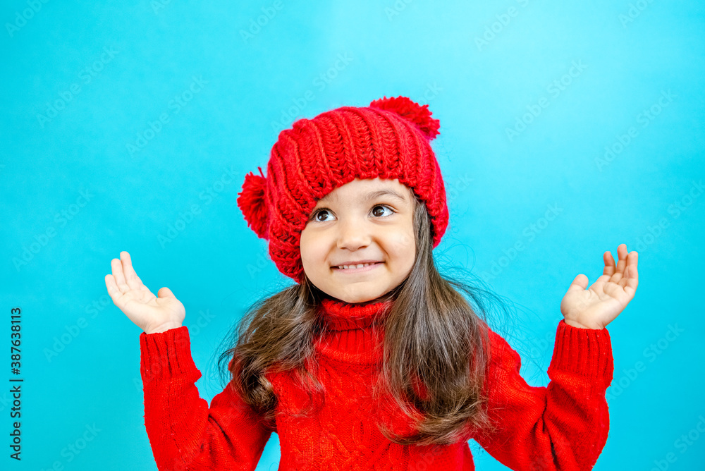 Portrait of a little curly-haired girl in a knitted red hat in winter. little girl with dark hair in a red knitted hat and sweater smiling on a blue background isolate, place for text. Winter clothes Portrait of a little curly-haired girl in a knitted red hat in winter. little girl with dark hair in a red knitted hat and sweater smiling on a blue background isolate, place for text. Winter clothes