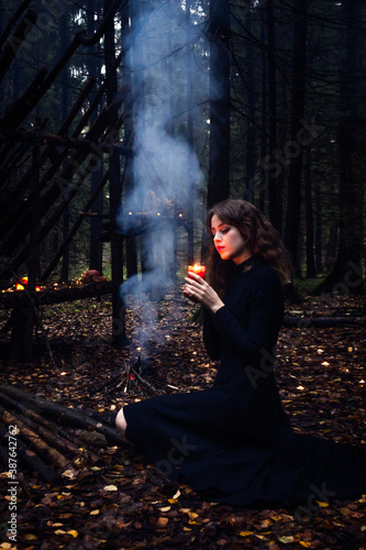 Halloween witch girl with witchcraft, near the fire, spells, in the forest. Beautiful young woman in conjuration. Spooky dark magic forest background. Wizard. Halloween party design