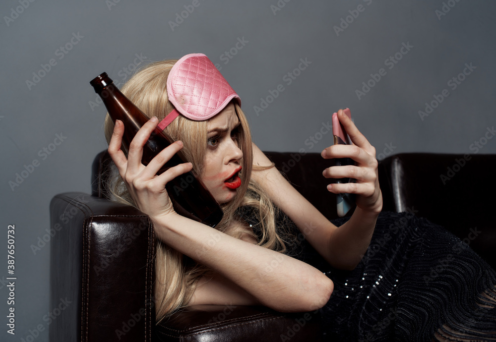 A drunk woman with a bottle of beer looks at a mobile phone in her hand and lies on the couch