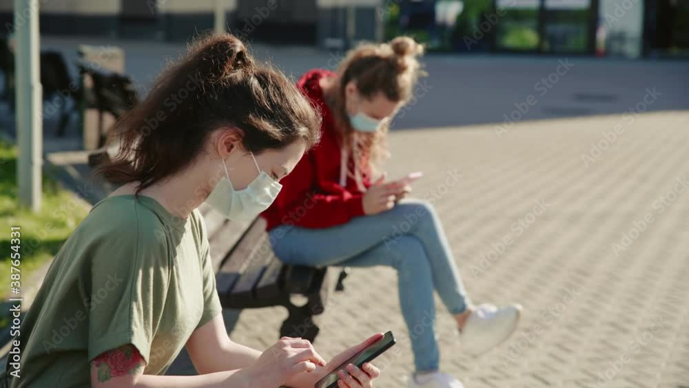Two young girls sitting on different benches far from each other in protective masks from the coronovirus. They taking precautions against covid-19 together. Isolation and social distancing concept. Two young girls sitting on different benches far from each other in protective masks from the coronovirus. They taking precautions against covid-19 together. Isolation and social distancing concept.