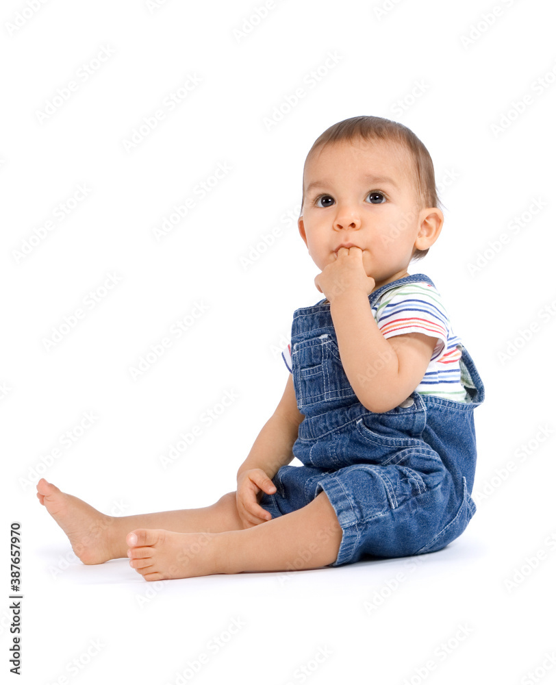 A baby boy sitting on the floor sucking his fingers, dressed in overalls and isolated on white background A baby boy sitting on the floor sucking his fingers, dressed in overalls and isolated on white background
