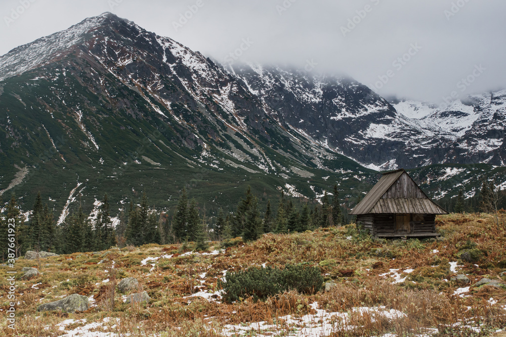 Old wooden cottage among high mountains, fiords landscape, with fog and snow all around. Concept of isolation, social distance travel, trekking in Europe. Moody, desaturated colors.