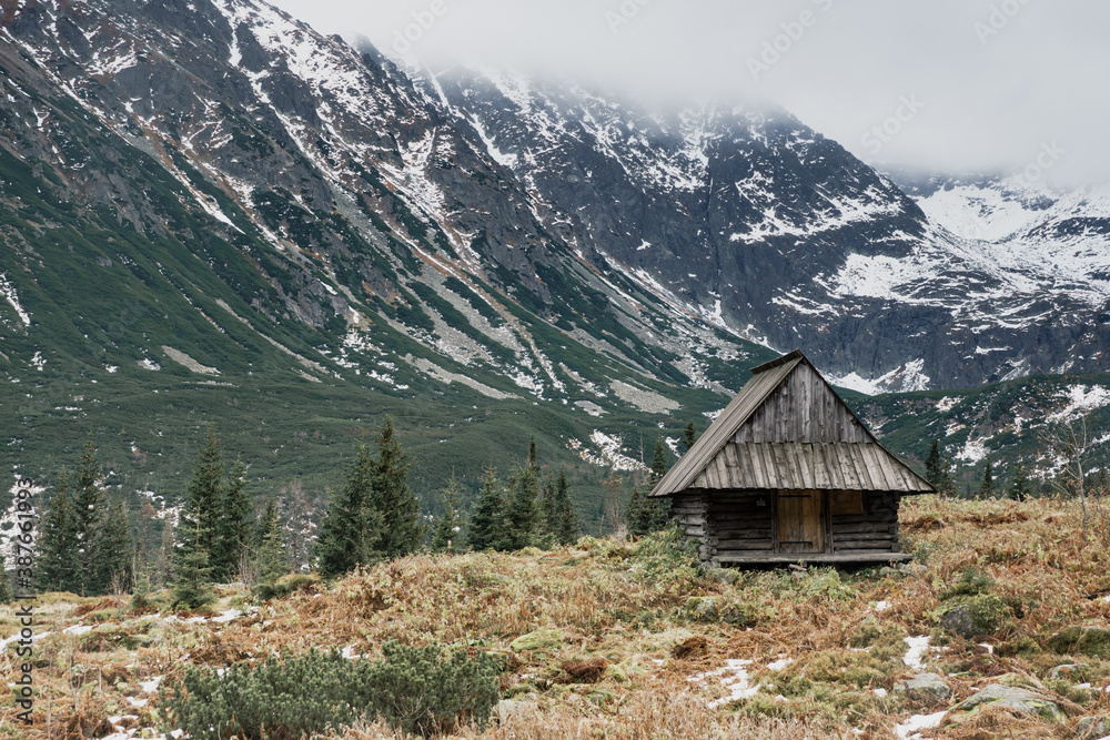 Old wooden cottage among high mountains, fiords landscape, with fog and snow all around. Concept of isolation, social distance travel, trekking in Europe. Moody, desaturated colors.