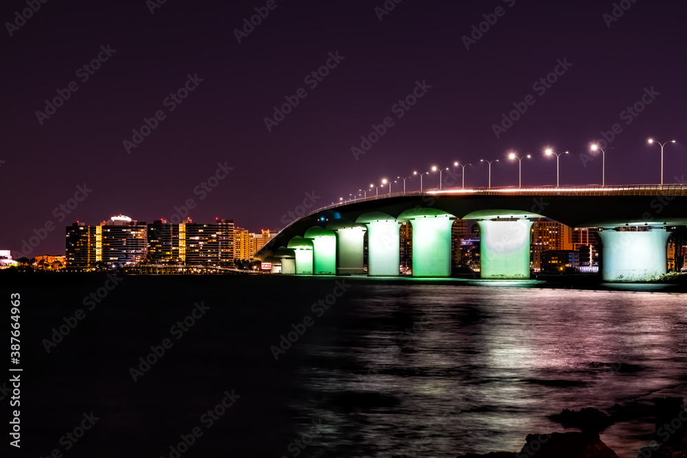 Naklejka premium John Ringling causeway bridge in Sarasota Bay Florida at dark night with city lights, skyscrapers buildings cityscape skyline in background and water reflection