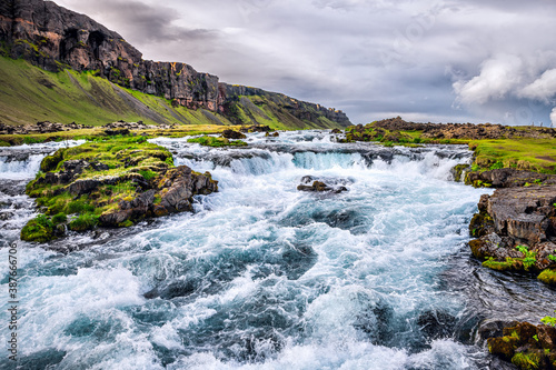 Landscape view of rushing river by Fjadrargljufur canyon in Iceland with rapids stream of water by green grass moss and cloudy sky