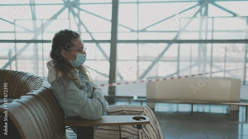 Young woman sitting alone in medical mask in airport at safe distance. female pasenger looking in smartphone waiting for her flight. New normal. Travel during pandemic. Post quarantine. photo