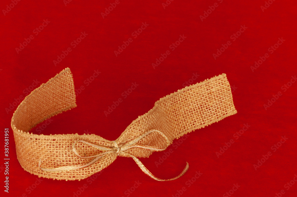 Fototapeta premium Ribbon at lower right corner framing text message space at top and right. Red background with burlap and gold ribbon bow in swirl curve shape.