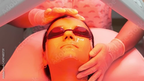 Young woman having red LED light facial therapy treatment in beauty salon. Hands of beautician adjusting collagen hydrogel mask on client face. Beauty, healthcare and wellness concept