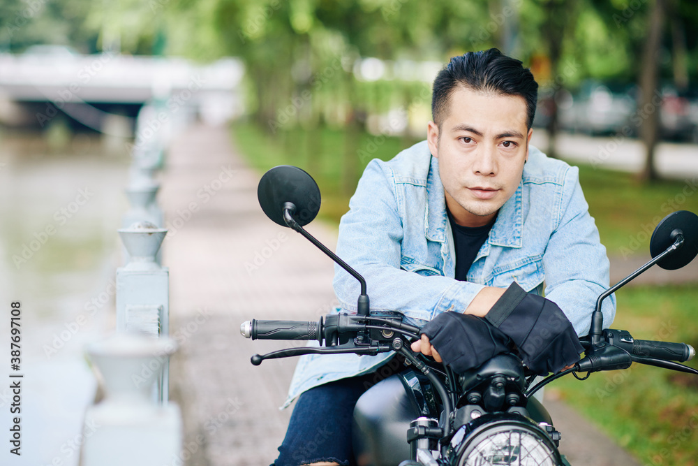 Portrait of serious handsome Vietnamese motorcyclist leaning on handlebar and looking at camera