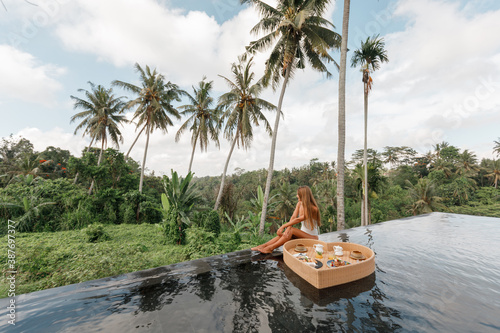  Pretty young woman enjoying floating breakfast in the pool at luxury villa, sit on the edge of infinity pool with coconut palm trees view