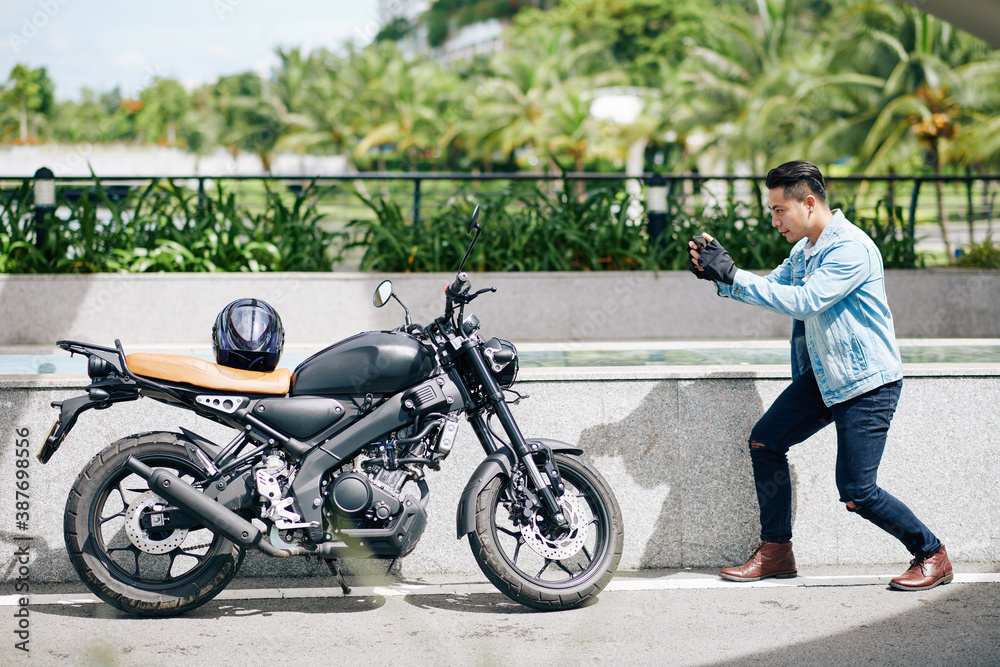 Handsome young Asian man taking photo of his motorcycle on smartphone