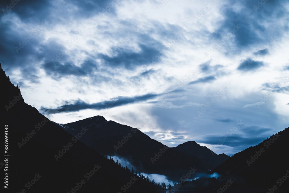 Low cloud above mountain lake. Mysterious silhouettes of big mountains and trees. Dense fog above firs and pines. Highland tranquil landscape at early morning. Ghostly alpine atmospheric scenery. Low cloud above mountain lake. Mysterious silhouettes of big mountains and trees. Dense fog above firs and pines. Highland tranquil landscape at early morning. Ghostly alpine atmospheric scenery.