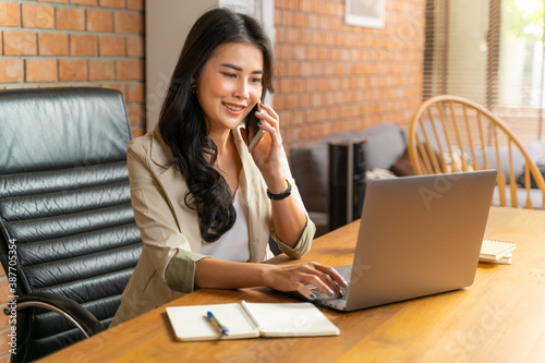 Happy young beautiful Asian business woman taking on the phone while using a computer during working from her home