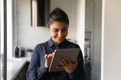 Happy millennial Indian woman look at modern tablet gadget screen browse surf wireless internet. Smiling young ethnic female texting messaging on pad device, use mobile computer. Technology concept.