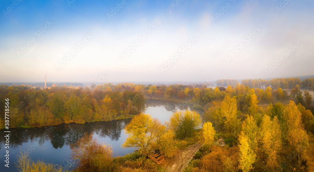 Fototapeta premium Autumn landscape: The river is surrounded by yellow trees in the fog in November. Warm calm panorama of autumn. 
