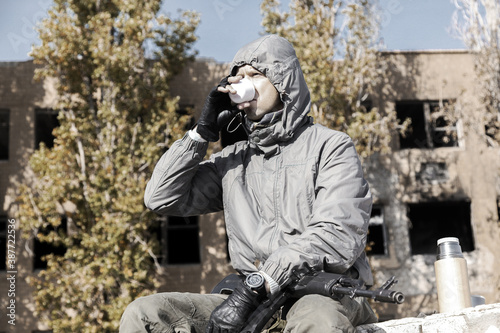 a confident male stalker in a jacket with a hood sits and drinks tea from a thermos, weapons on his lap, against the background of a ruined house