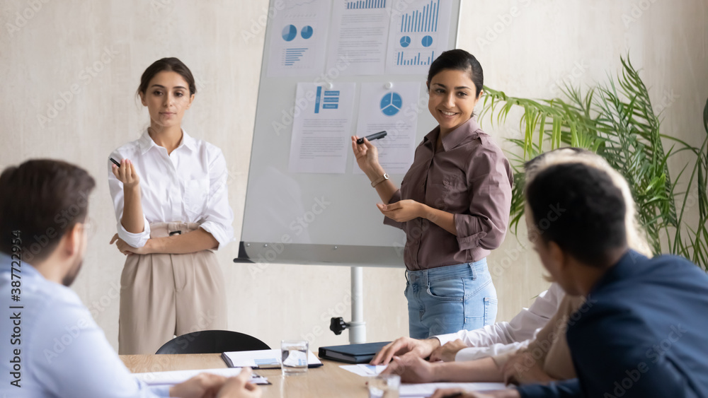 Smiling multiracial female speakers talk interact with employees make whiteboard presentation at office meeting. Multiethnic trainers present business project startup on flip chart at group briefing.