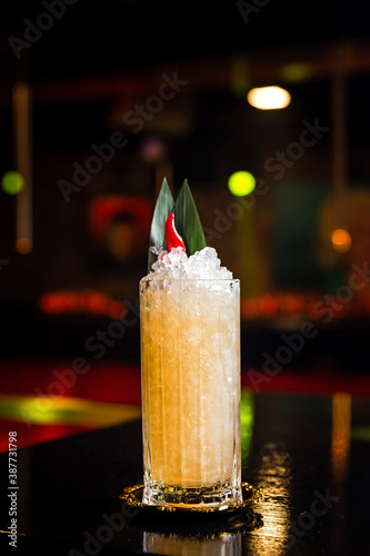 An orange cocktail in a collins highball glass on crushed ice, garnished with banana leaves and red pepper