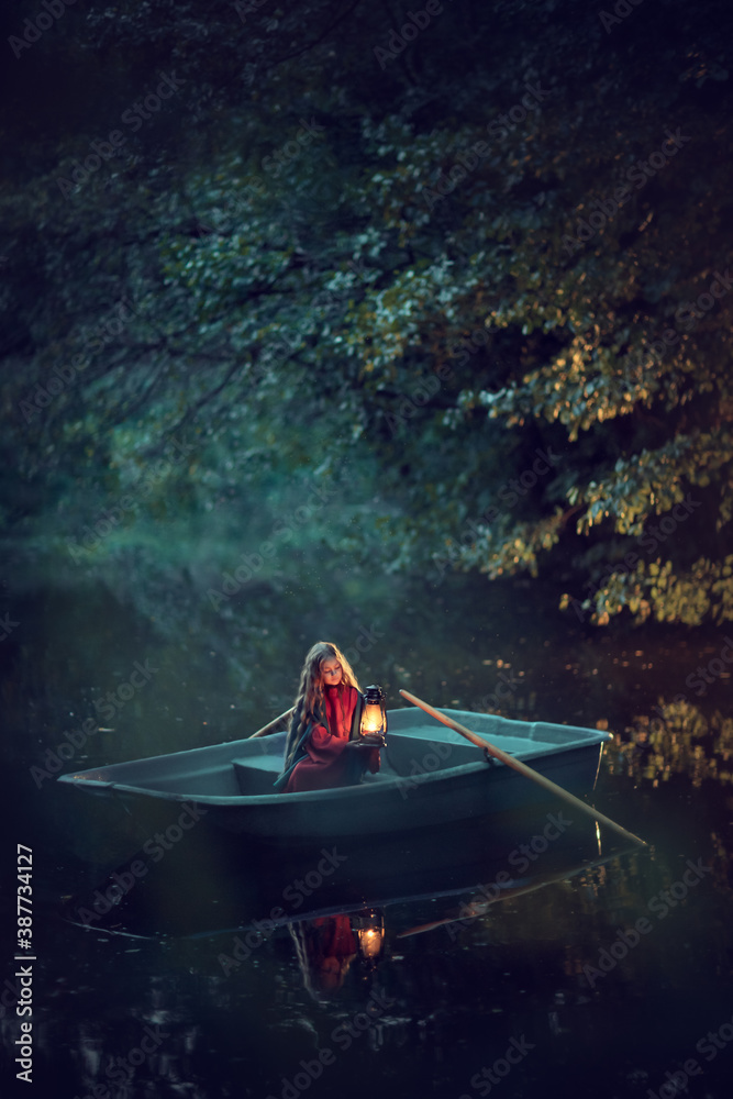 Naklejka Beautiful calm girl in the cape in the boat with the lantern in the dark forest river. Night magical wood
