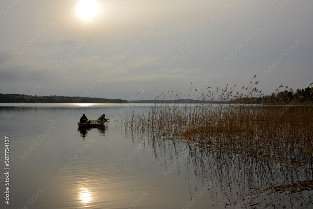 Two fishermen fishing from a boat in lake center. Sun shining from misty clouds and reflect in water.
