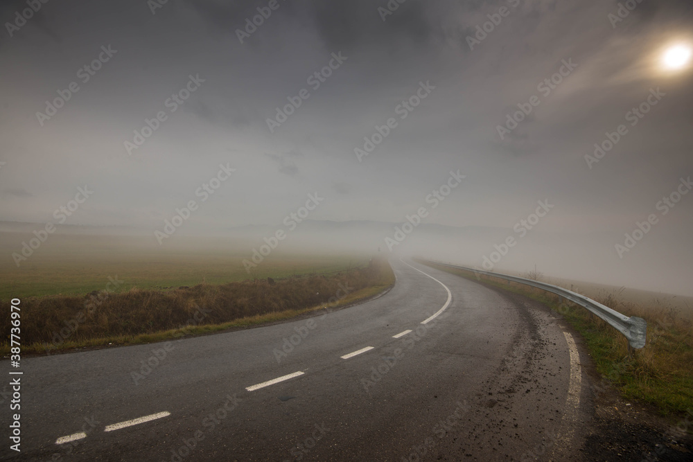 Fototapeta premium Curving, wet asphalt road in dense fog, sun shining behind thick clouds.