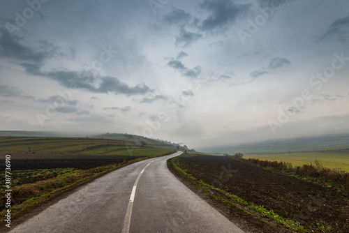Wet asphalt road leading through green agricultural fields at late autumn, fog and thick rain clouds.