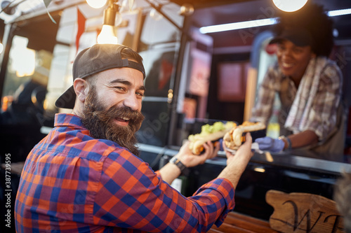 happy, satisfied caucasian beardy male customer taking  sandwiches from a polite employee in fast food service, looking at camera, laughing © luckybusiness