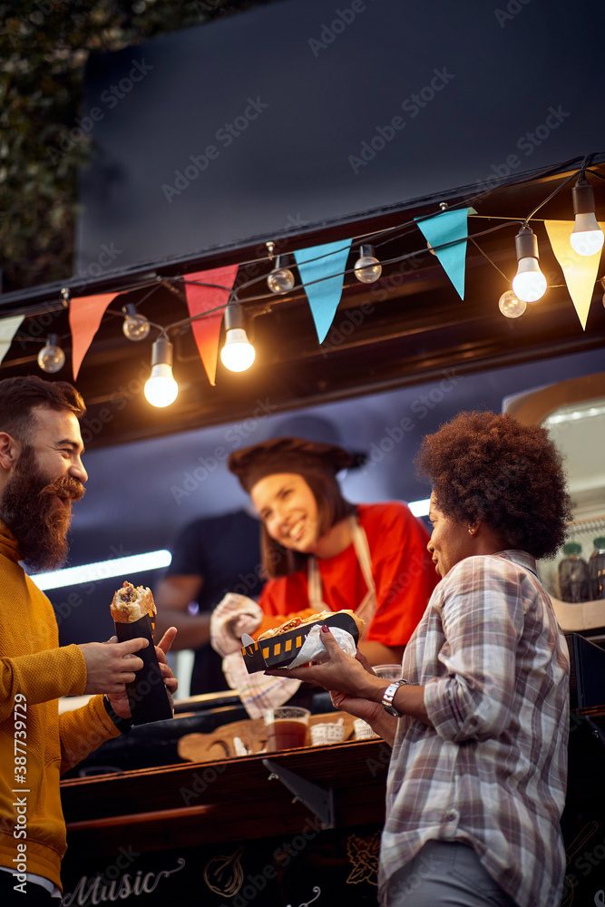 multiethnic couple having a good time, talking, eating sandwiches outdoor in front of fast food object