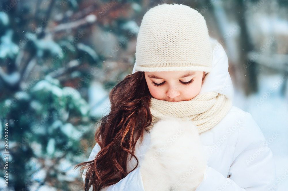 Winter close up portrait of cute dreamy child girl in white coat, hat and mittens playing outdoor in snowy winter forest. Traveling, vacation and active winter holidays concept