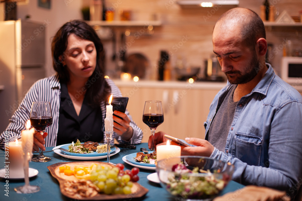 Couple holding glasses of wine and using smartphone in kitchen during romantic dinner with candles and food in the table. Adults sitting at the table browsing, searching, using smartphones, internet,