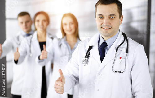 Group of professional doctors are standing as a team with thumbs up in a sunny hospital office  ready to help their patients. Medical help  insurance in health care and medicine concept