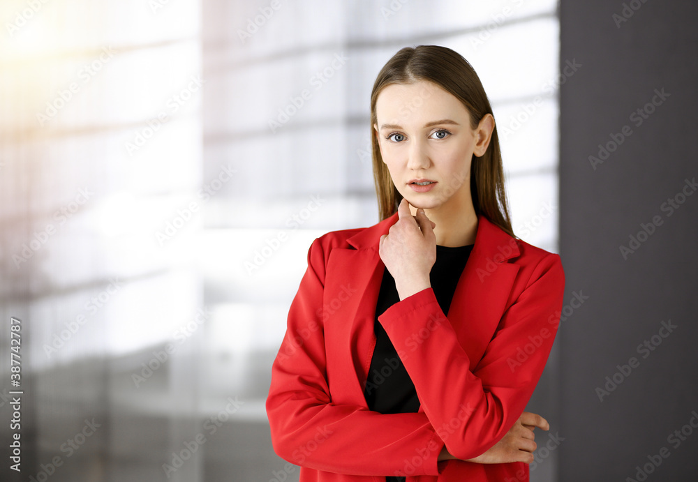 Friendly young business woman or female student dressed in red coat is standing straight and looking at camera in a sunny office. Urban lifestyle and diverse people concept Friendly young business woman or female student dressed in red coat is standing straight and looking at camera in a sunny office. Urban lifestyle and diverse people concept