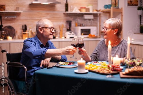 Handicapped man having dinner with man in kitchen toasting. Wheelchair immobilized paralyzed handicapped man dining with wife at home  enjoying the meal