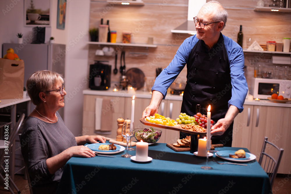 Senior man serving grapes his wife during romantic dinner in kitchen for relationship anniversary Elderly old couple talking, sitting at the dining table enjoying the meal, Senior man serving grapes his wife during romantic dinner in kitchen for relationship anniversary Elderly old couple talking, sitting at the dining table enjoying the meal,
