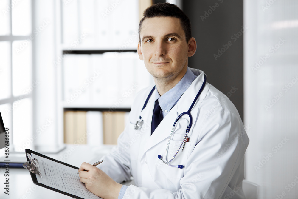 Friendly male doctor sitting and working with clipboard of medication history records in clinic at his working place. Perfect medical service in hospital. Medicine and healthcare concept